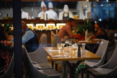 empty served table in an expensive restaurant for four people with glasses, plates and appliances in the background visitors and 3 cooks in white. evening interior of a popular cafe clipart