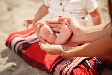 Manikürlü genç bir anne elleriyle küçük ayaklarını tutuyor ve kumsalda ayaklarını bir ekosenin üzerinde gösteriyor. Yüzü olmadan yakın çekim, deniz kenarında piknik, ailesiyle tatil.