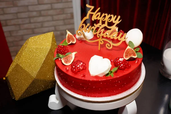 Delicious red cake with sparkles for a birthday with a large golden diamond for a young woman, decorated with halves of pomegranate and passion fruit. Wooden inscription happy birthday on a stick.