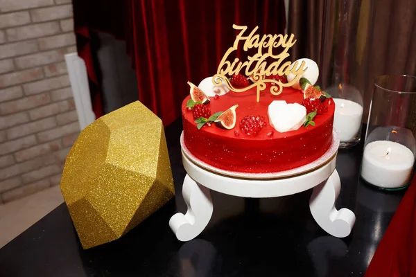 Delicious red cake with sparkles for a birthday with a large golden diamond for a young woman, decorated with halves of pomegranate and passion fruit. Wooden inscription happy birthday on a stick.