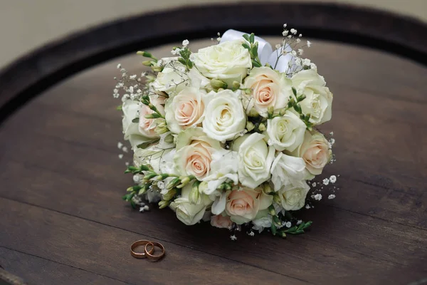 A wedding bouquet with lively white and pink roses lies on a wooden barrel near the gold wedding rings. Symbols of eternal love on the table. Natural bouquet of beautiful flowers. Accessory bride at the wedding
