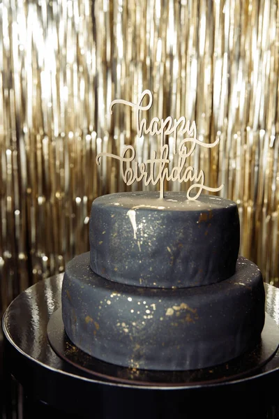 大きな黒い誕生日ケーキ金のスパンコールの背景に光沢のあるテーブルの上で誕生日の碑文と金の滴 — ストック写真