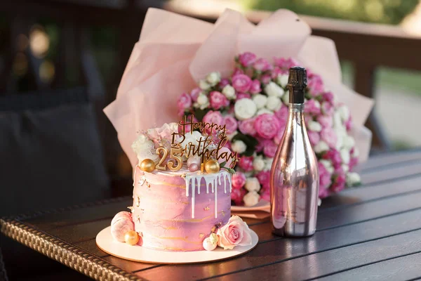 木製の台の上に金色の数字の美しい層状のピンクのケーキバラの大きな花束と光沢のあるボトルのシャンパンのボトルの近くの若い女の子のための25歳の誕生日 — ストック写真