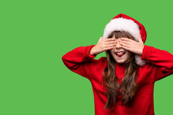 戴着圣诞老人帽的女孩用手捂住眼睛 伸出舌头 一个被绿色背景隔离的孩子的情感画像 新年气氛 高质量的照片 — 图库照片