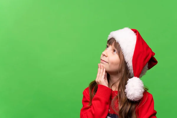 一个穿着红色针织毛衣 头戴圣诞帽的快乐的小女孩若有所思地看着我 梦想着礼物 许下了一个愿望 圣诞快乐 新年快乐 高质量的照片 — 图库照片