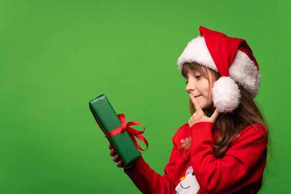 一个戴着圣诞礼帽的小女孩手里拿着圣诞礼物 试图用猜盒子里的东西 被绿色背景隔离 高质量的照片 — 图库照片
