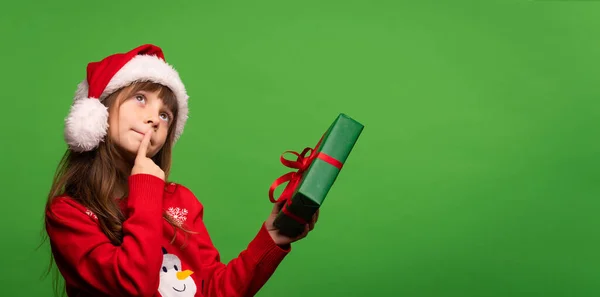 一个戴着帽子的可爱的圣诞老人小女孩手里拿着一份新年礼物 梦游般地抬起头 试图用猜盒子里的惊喜 圣诞快乐 新年快乐 被绿色背景隔离 — 图库照片