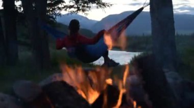 Kamp ateşinin yanındaki hamakta dinlenen genç bir kadın Kanada, Alberta, Yaha Tinda 'daki Eagle Creek Kampı' ndaki Kızıl Geyik Nehri 'ne bakıyor..