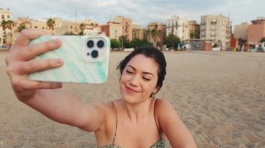 Genç bir kadın kumsalda oturuyor ve cep telefonu kullanıyor. Video görüşmesi yapıyor.