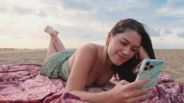 Kumsalda dinlenen ve cep telefonu kullanan gülümseyen genç bir kadın.