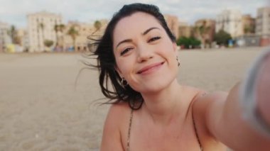 Kapalı, gülümseyen bir kadın kumsalda oturuyor ve cep telefonu kullanıyor. Video görüşmesi yapıyor.