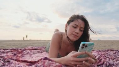 Genç bir kadın sahilde dinleniyor ve cep telefonu kullanıyor.
