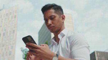 Kapatın, genç işadamı iş merkezinin yanında otururken cep telefonundan bilgileri inceliyor.