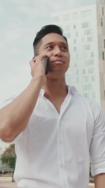 Genç işadamı, modern binaların arka planında akıllı telefondan konuşuyor.