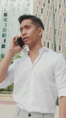 Genç işadamı, modern binaların arka planında akıllı telefondan konuşuyor.