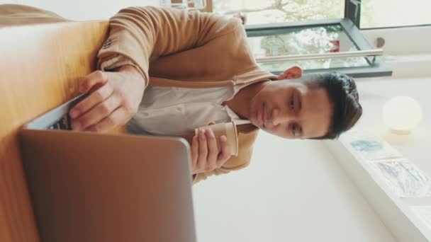 Vertical Video 身穿米色西服的年轻英俊商人在咖啡店里和笔记本电脑一起工作 手里拿着一杯咖啡 — 图库视频影像