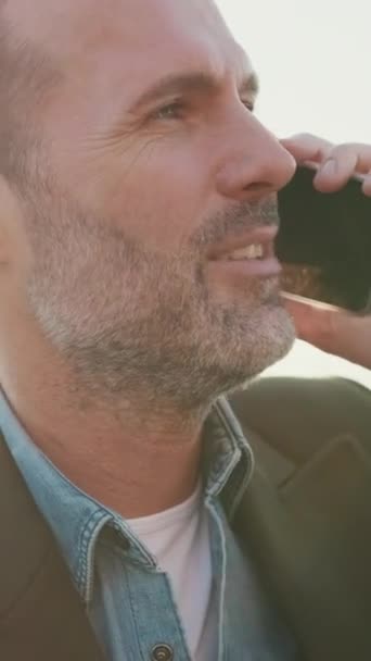 垂直方向のビデオ 閉じると 男は携帯電話で話しながら 川の堤防に座っている バックライト — ストック動画