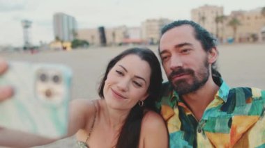 Sahilde otururken cep telefonuyla selfie çeken romantik çiftin yakın çekimi.