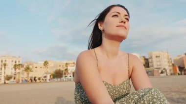 Genç bir kadın kumsalda oturuyor ve uzaklara bakıyor.