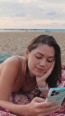 Genç bir kadın kumsalda dinlenirken akıllı telefon kullanıyormuş, bu doğru mu? Kız başını kaldırır ve kameraya bakıp gülümser.