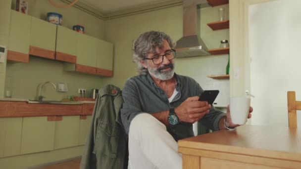 キッチンに座っている間 彼のスマートフォンや飲み物のコーヒーに幸せなシニア読書メッセージ — ストック動画