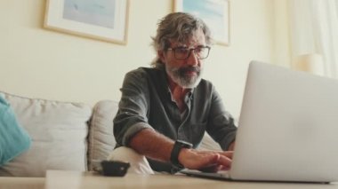 Oturma odasındaki kanepede oturan, dizüstü bilgisayarda yazan yaşlı pozitif bir adam.