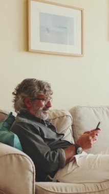Kanepede oturan, cep telefonu kullanan, evdeki oturma odasında dinlenen yaşlı bir adam.