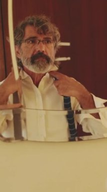 Orta yaşlı bir adam aynanın önünde dikilirken kravat takıyor.