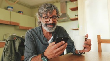 Yaşlı adam fincandan kahve içiyor ve evde cep telefonu kullanıyor.