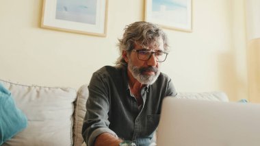 Oturma odasındaki kanepede oturan, dizüstü bilgisayarda yazan yaşlı pozitif bir adam.