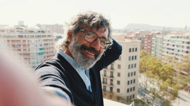 Yetişkin bir adamın balkonda dikilirken selfie çektiği yakın plan çekimde kameraya bakar.