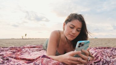 Kumsalda dinlenen ve cep telefonu kullanan gülümseyen genç bir kadın.