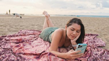 Kumsalda dinlenen ve cep telefonu kullanan gülümseyen genç bir kadın.