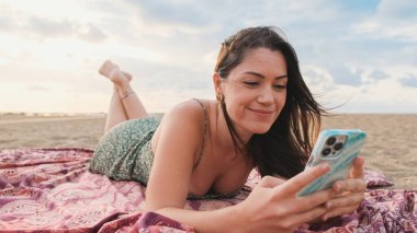 Genç bir kadın sahilde dinleniyor ve cep telefonu kullanıyor.