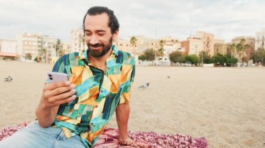 Gülümseyen adam elinde akıllı telefonuyla kumsalda oturup rahatlıyor.