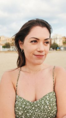 Genç kadın sahilde duruyor ve uzaklara bakıyor. Kız başını çevirir, kameraya bakar ve gülümser.