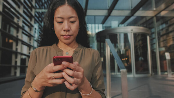 Close-up of young happy businesswoman using mobile phone while standing on modern buildings background