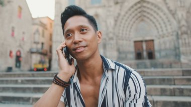 Genç turist merdivenlerde oturuyor. Arka planda manzaralı cep telefonuyla konuşuyor.