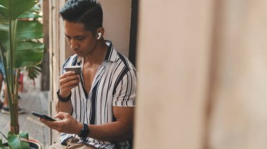 Genç adam dışarıda oturup kahve içiyor ve cep telefonu kullanıyor.