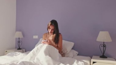 Sabah uyandıktan sonra telefonda mesaj atan mutlu kadın. Gülümseyen kız internette konuşuyor, evde yatakta otururken mesajlarına cevap veriyor.