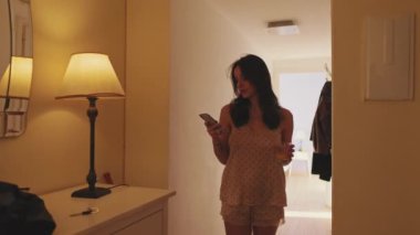 Pijamalı genç kadın camdan meyve suyu içiyor ve yatak odasından çıkarken cep telefonu kullanıyor.