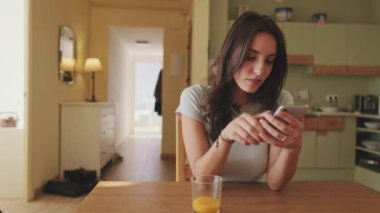 Genç kadın portakal suyu içiyor ve mutfakta otururken akıllı telefon kullanıyor.