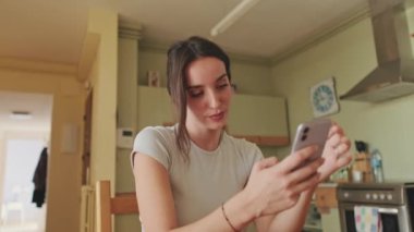 Genç bir kadın mutfakta otururken cep telefonunu kullanıyor.