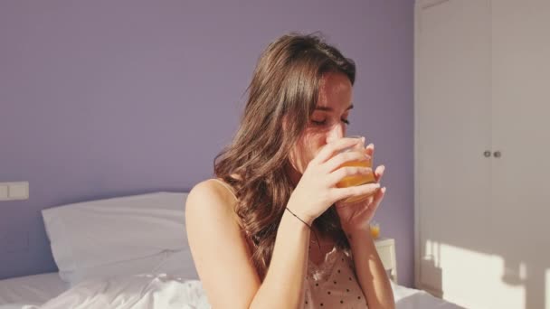 寝室のベッドに座っている間 パジャマ姿の若い女性がガラスからジュースを飲む 女の子は窓の外を見て楽しい晴れた日 — ストック動画