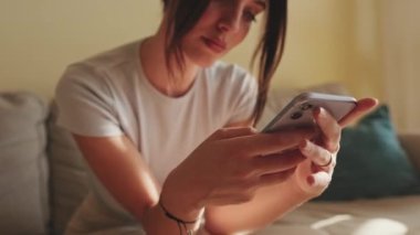 Evdeki kanepede otururken genç bir kadının cep telefonunu mesaj atmak ve kaydırmak için kullanması.