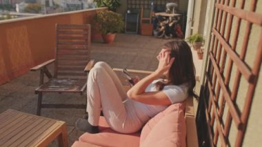 Evde balkonda otururken cep telefonu kullanan genç bir kadın.