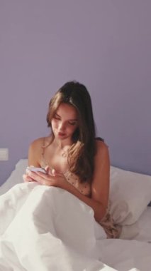 VERTICAL VIDEO, Mutlu kadın sabah uyandıktan sonra telefonla mesaj atıyor. Gülümseyen kız internette konuşuyor, evde yatakta otururken mesajlarına cevap veriyor.