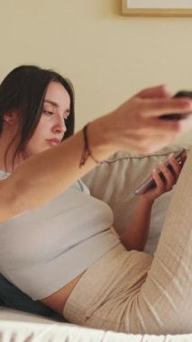 VERTICAL VIDEO, evdeki kanepede otururken uzaktan kumandayla kanal değiştiren genç kadın