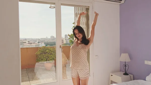 Young woman in pajamas stretches when standing by the window in the bedroom