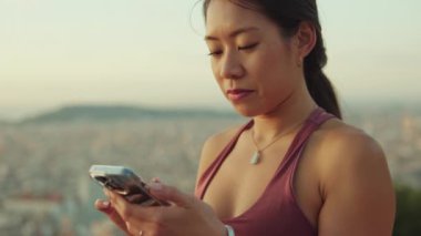 Clopse up, happy athletic woman using mobile phone while standing at lookout point at dawn